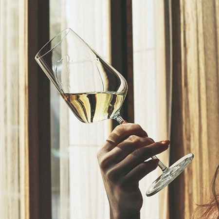 Hand hält ein Glas Weißwein am Fenster