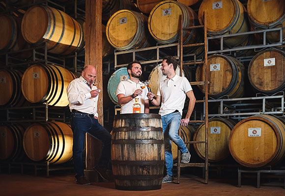Drei Männer verkosten Spirituosen vor Holzfässern in einer Brennerei