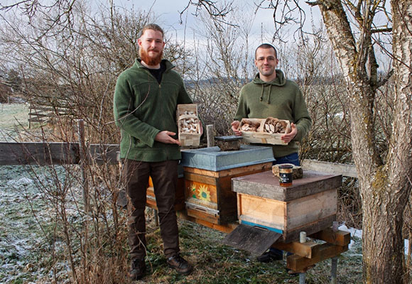 Zwei Imker stehen neben Bienenstöcken und zeigen Produkte der Wildnismanufaktur