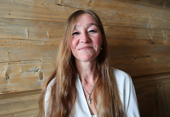 Portrait einer Mitarbeiterin mit langen Haaren und Kreuzkette vor Holzwand