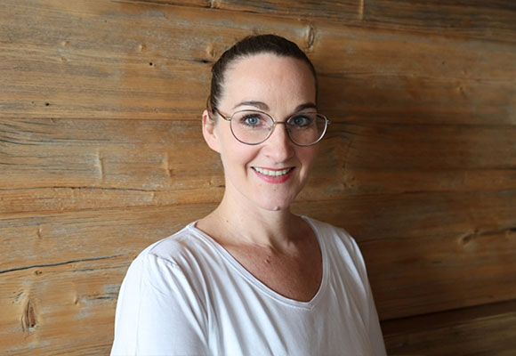 Mitarbeiterin mit Brille und zusammengebundenen Haaren lächelt vor Holzwand