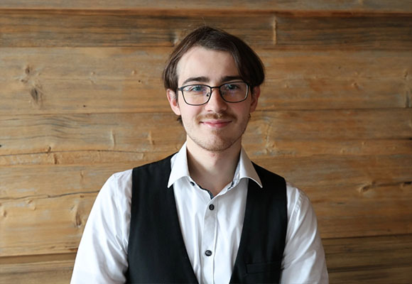 Junger Mitarbeiter mit Brille und Weste vor Holzwand