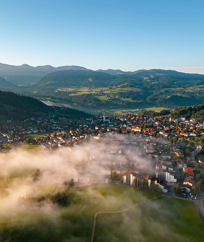Luftaufnahme von Oberstaufen mit Nebel über der Ortschaft und Berglandschaft