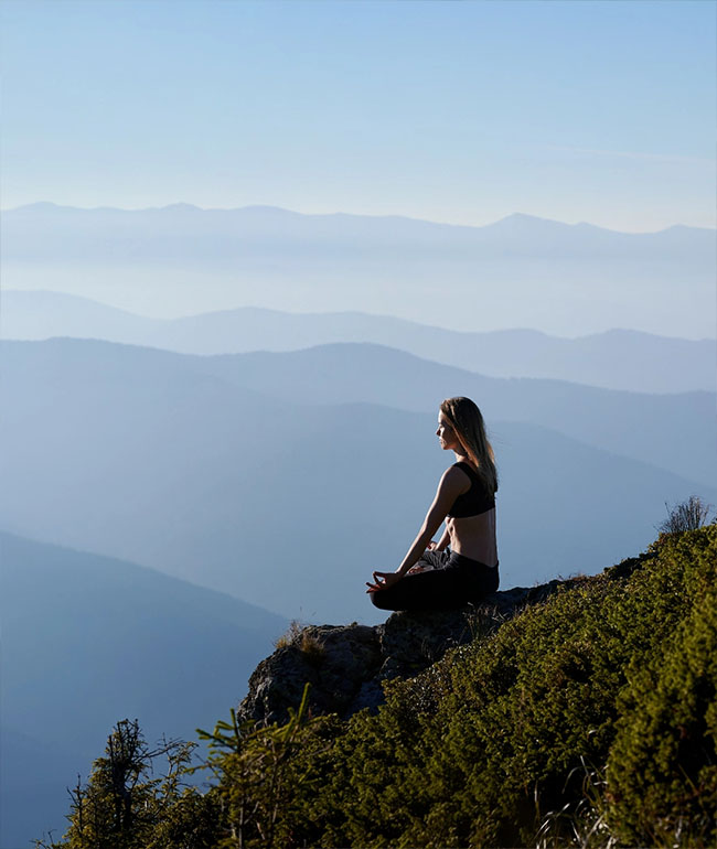 Frau sitzt meditierend auf einem Berg mit Blick über die Landschaft