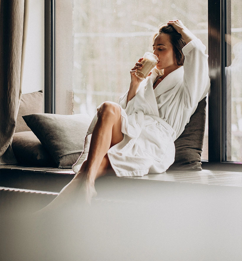 Frau im Bademantel sitzt entspannt am Fenster und trinkt Kaffee